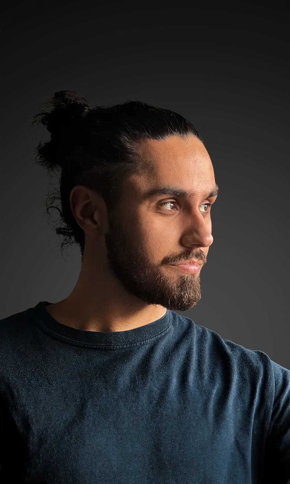 Portrait of a man with dark hair in a messy bun, beard, and t-shirt, looking to the side